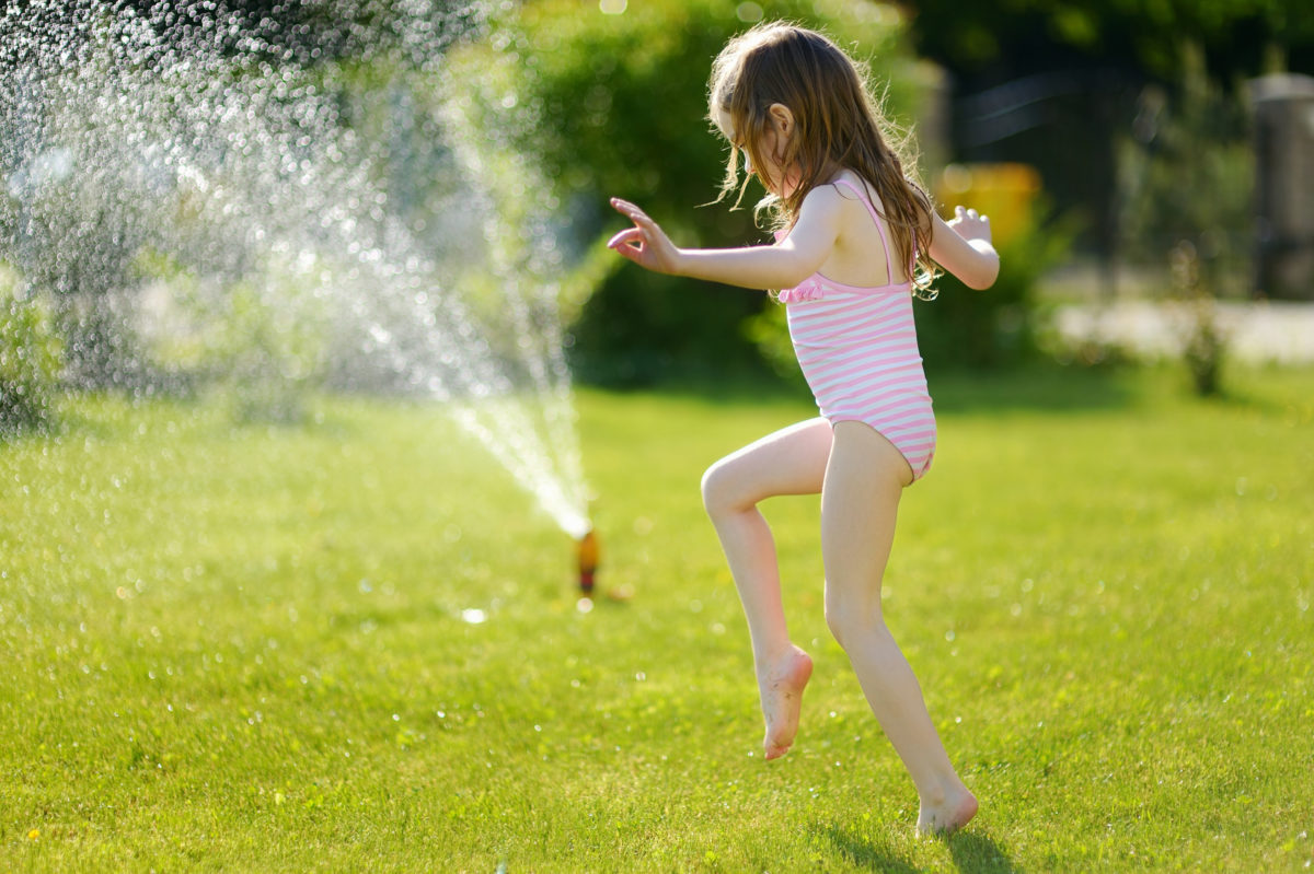 Girl Running Though A Sprinkler In A Backyard Natural Tree